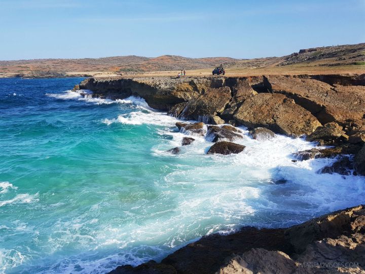 Imagem de uma paisagem mais desconhecida de Aruba, com o mar bem agitado, de um azul esverdeado bem forte, muitas pedras e um jipe